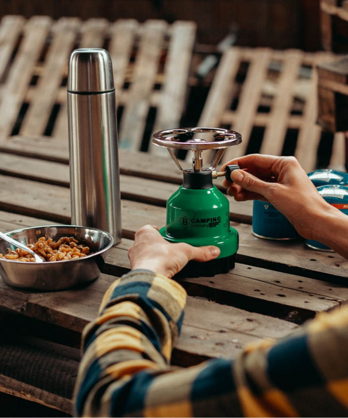 Auf Holzpletten steht ein Campingkocher, Gaskartuschen sowie eine Thermusflasche und eine Metalchalte mit Essen drinnen. Eine Person drhet am Gaskocher.