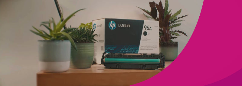 Auf einem Schreibtisch stehen Pflanzen sowie eine Schachtel eines HP Toners. Eine Person erscheint im Bild und stellt eine Schchtel eines Canon Toners hinzu.